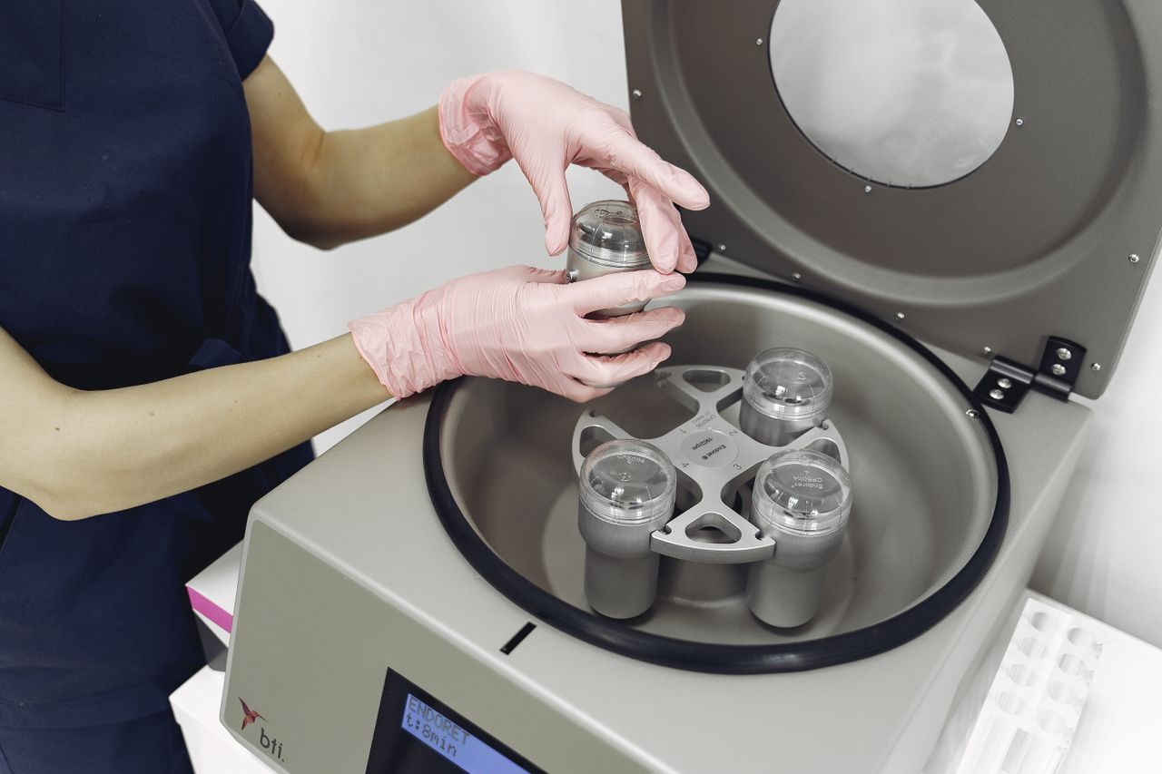 From above of crop female nurse in uniform and latex gloves standing near centrifuge and preparing equipment for plasmolifting procedure in salon